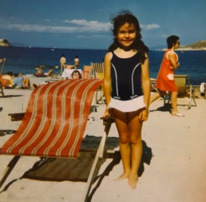 Young Julie Graham on the beach in her costume and posing next to a deckchair