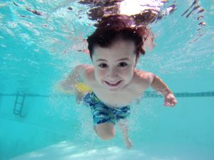 Little boy swimming underwater in a pool with a big smile on his face.