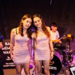 2 teen girls in pink dresses singing into a microphone with a boy playing drums in the background at Wejam